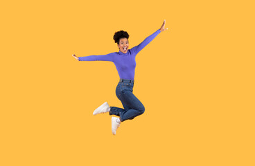 Obraz premium A young woman dressed in a purple top and blue jeans jumps exuberantly, displaying a wide smile. The bright yellow backdrop adds vibrancy, capturing a sense of happiness and energy