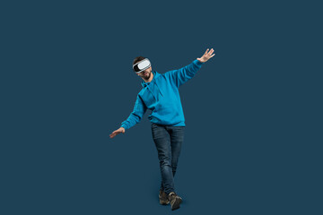 A young man engages in a balance exercise while wearing a virtual reality headset. He stands on one leg, arms outstretched for stability, in a modern indoor setting with a solid color backdrop