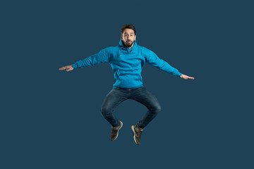 Obraz premium A young man demonstrates his enthusiasm by leaping off the ground in a bright blue hoodie, showcasing his playful spirit and joy. The solid dark background emphasizes his energetic pose