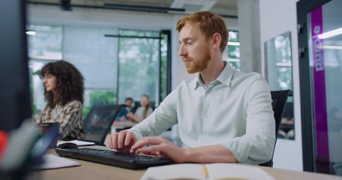 Caucasian man with stylish shirt and red hair working in busy office. Typing on keyboard. Writing code or message to someone. Noting something down on small notepad for later. Busy hour. Workplace.