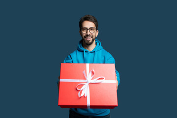 A cheerful young man wearing a blue hoodie stands with a vibrant red gift box in his hands. His bright smile and stylish glasses create a warm atmosphere