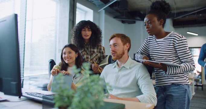Multi-ethnic group of people discussing something in front of monitor at big desk. Man with beard placing notepad with some valuable information on table. Everyone smiling. Team building activity.