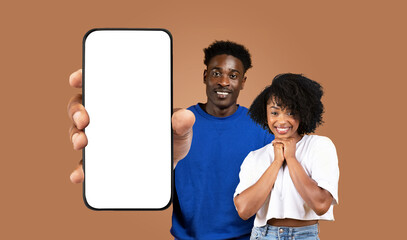 African American young man and woman stand in front of a brown background, the man holding a smartphone with a blank screen in front of him. Both are smiling directly at the camera