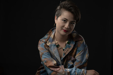 Young woman wearing a stylish shirt, smiles subtly while looking pensively, against a dark background, exuding thoughtfulness and modern style with a unique necklace.