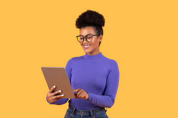 A young woman with curly hair is smiling while using a tablet, showcasing her engagement and enjoyment with the device. She wears stylish glasses and a long-sleeved purple top