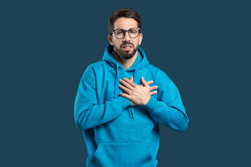 A young man in a blue hoodie stands with his hands on his chest, displaying a deep expression of emotion. The dark background highlights his thoughtful demeanor