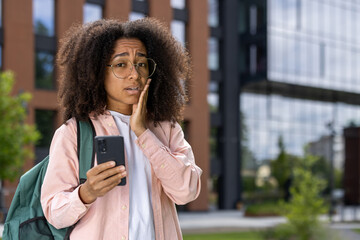 African American female student stands outdoors, looking concerned while holding a smartphone. The...
