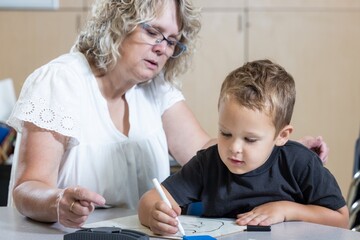 A young boy is sitting at a table with a teacher, writing with markers. The teacher is watching the boy and seems to be helping him with his work. The scene is a learning environment for world teacher