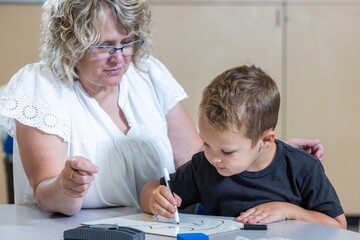 A young boy is sitting at a table with a teacher, writing with markers. The teacher is watching the...