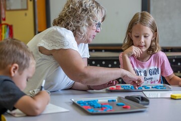 A teacher is helping a young girl with a spelling. The teacher is pointing to a letter, and the girl is looking at it intently.