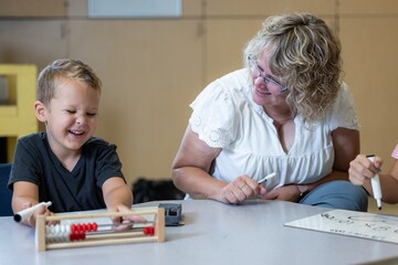 A young boy is sitting at a table with an abacus and smiling with his teacher laughing with him as he solves the problem.