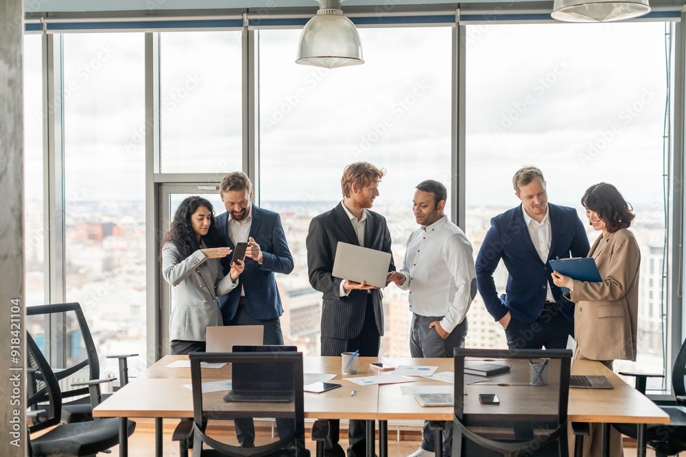 Poster A group of business professionals stand around a large table in a modern office. There is a view of a city through the large windows behind them.