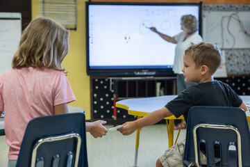 A boy and a girl are sitting at a table in a classroom passing notes to each other. The girl is wearing a pink shirt and the boy is wearing a black shirt. A teacher is standing in front of a whiteboar