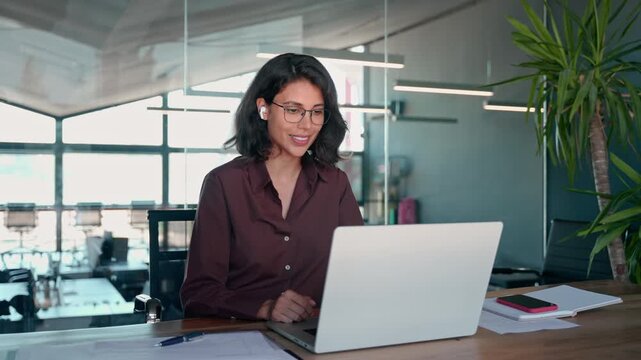 Middle easter business woman in earphones having virtual team meeting video conference call. Latin businesswoman using laptop talking to coworkers, partners in remote online chat, sitting in office