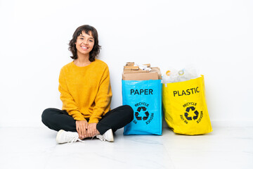 Young girl holding a bag full of plastic and paper sitting on the floor isolated on white background posing with arms at hip and smiling