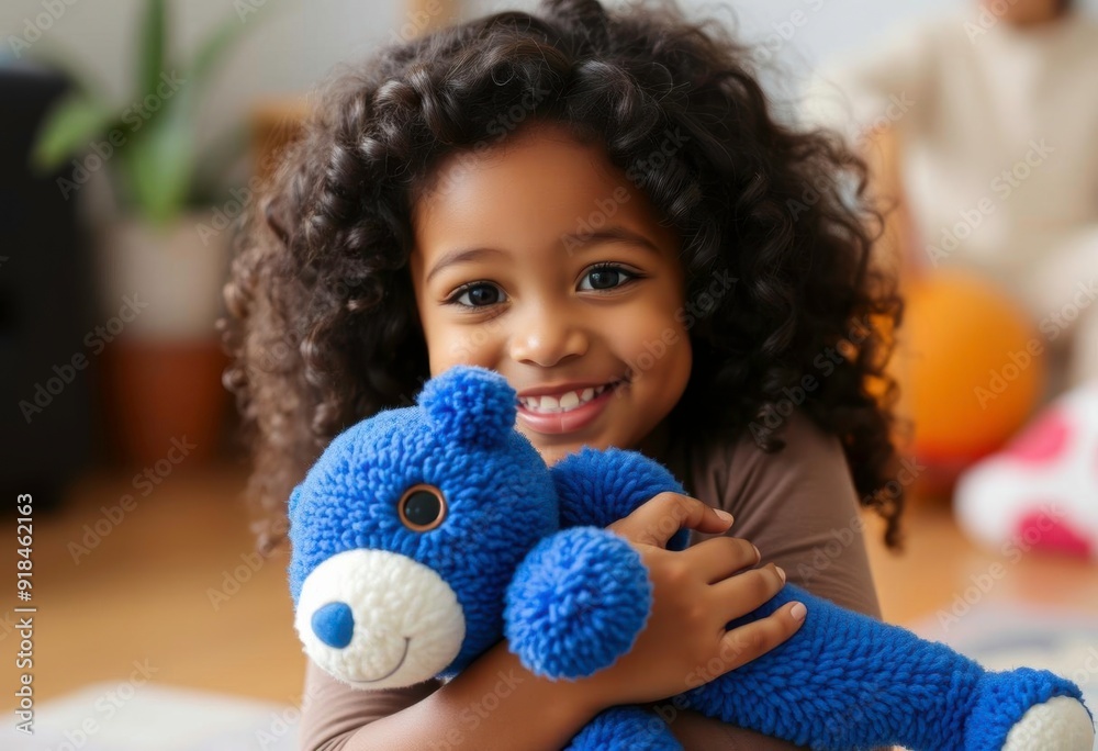 Wall mural A young African American girl with curly dark hair, smiling and holding a blue stuffed animal