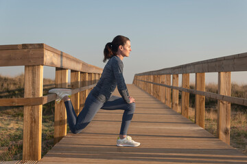 Sporty mid adult woman doing a leg stretch warm up exercise outdoors on a wooden boardwalk