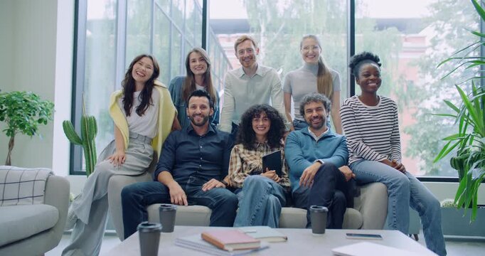 Camera moving closer to multi-ethnic group of friends. Everyone gathering together next to one sofa in lounge area. Engineers or programmers posing for camera in big room. Sunny weather in background.