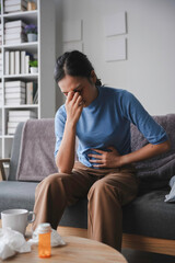 Young woman sitting on a sofa, holding her stomach and feeling sick, with medicines and tissues on...