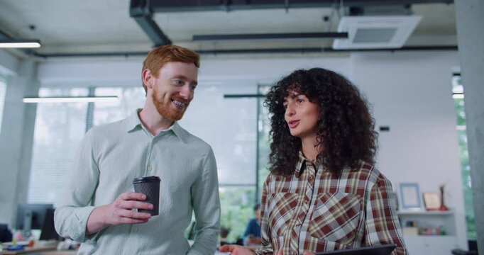Camera view of two Caucasian people talking in office. Handsome man holding cup of coffee in his hands while woman carrying tablet device. Workers with interest speaking and smiling to each other.