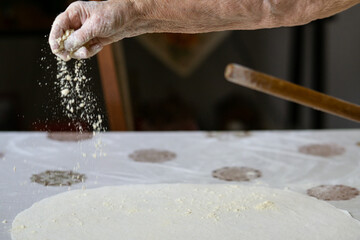Old woman hand pouring flour on a raw dough while rolling it with a rolling pin