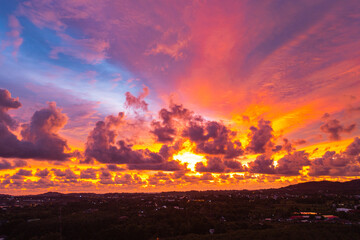 A breathtaking aerial view capturing a vibrant sunset over the Phuket countryside. The sky is ablaze with rich hues of yellow, orange, pink, and purple, while dramatic clouds float above the city
