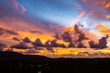 A breathtaking aerial view capturing a vibrant sunset over the Phuket countryside. The sky is ablaze with rich hues of yellow, orange, pink, and purple, while dramatic clouds float above the city