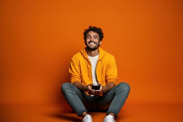 Full body young smiling happy Indian man he wears t-shirt casual clothes sit hold in hand use mobile cell phone look aside isolated on orange red color background studio portrait