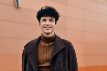 Fashion portrait of a young man with curly hair standing in front of an orange wall, smiling and wearing a brown coat and turtleneck