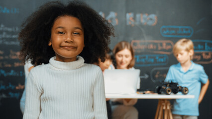 African girl smiling and looking at camera while multicultural friends working or learning engineering code or prompt in STEM technology classroom from young beautiful caucasian teacher. Erudition.