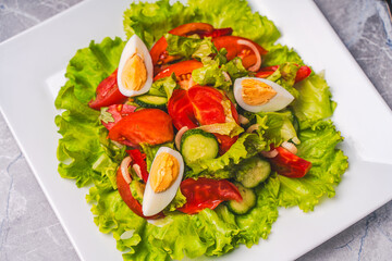 Vegetable salad of tomatoes, cucumbers, lettuce, red peppers and onions with boiled eggs on a white plate.