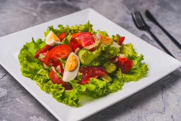 A light vegetable salad for healthy dietary nutrition. Tomatoes, cucumbers and boiled eggs with lettuce and traditional French vinaigrette sauce, on a white plate on a gray table.