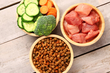 Bowls with dry pet food, raw meat and natural products on light wooden background, closeup