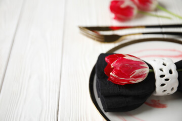 Plate with folded napkin, gold cutlery and tulip flowers on white wooden background