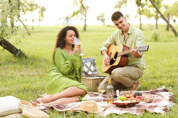 Happy boyfriend playing guitar for his girlfriend with glass of wine on romantic picnic in park