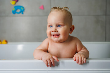 A smiling baby is standing in a white bath with water. Baby bathing in water. Personal hygiene of the baby.