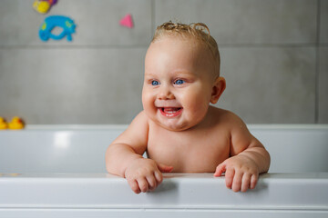 A smiling baby is standing in a white bath with water. Baby bathing in water. Personal hygiene of the baby.