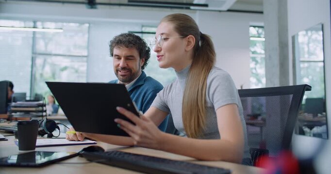 Intelligent Caucasian employees sitting together at table and discussing about new project. Handsome man with coworker comparing data from clipboards. Entering important information on computer.