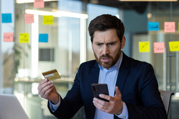 Concerned businessman in suit holding credit card and phone, looking at phone screen with worried expression, dealing with online banking or payment problem in modern office setting