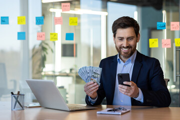 Businessman in suit smiling while holding cash and using smartphone at office desk. Man working on laptop, making online transactions, managing finances. Concept of success, financial growth, business