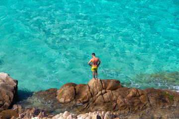 Mann auf Felsen in Badehose vor t&uuml;rkisem Meer