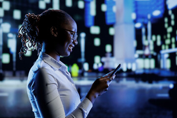 BIPOC woman walking around city at night, scrolling on online shopping platform using mobile phone....