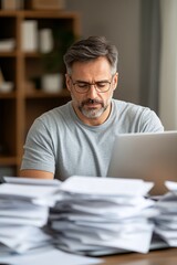 Obraz premium Middle-aged Man Working on Laptop Amidst Stacks of Papers at Home Office