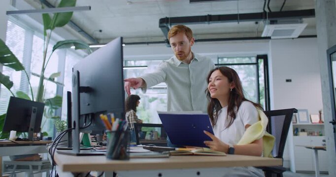 Smart leader standing behind woman and helping her with work at computer. Pretty woman sitting at table and holding folder with documents. Man showing thumbs up to female. Productive teamwork.