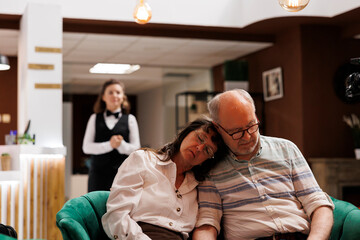 Comfortable hotel lobby with caucasian elderly couple relaxing on comfy couch. Photo showing exhausted retired old man and woman sleeping on sofa in modern lounge area, waiting for check-in.