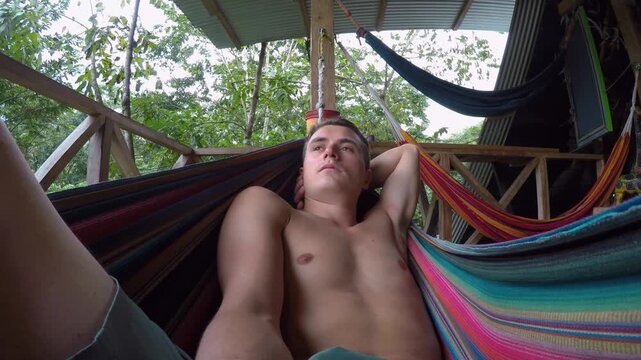 A shirtless young man lays on a colorful hammock at a hostel