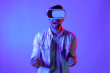 Happy excited man watching at sport competition at virtual world while wearing VR headset at neon light background. Handsome teenager enter future innovation technology by using goggles. Deviation.