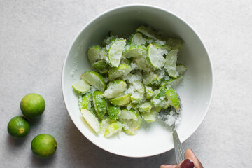Overhead view of lime slices and sugar being mixed together in a white bowl, process of making lime cheong or lime syrup