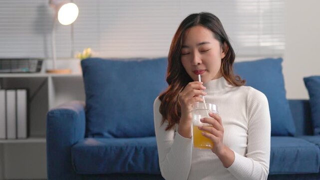Happy young woman enjoys a refreshing drink at home, smiling and laughing on a cozy blue sofa. She radiates joy and relaxation in her casual indoor setting