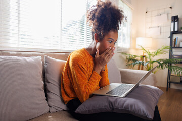 Image of cheerful excited happy african american woman afro hair,  using laptop while sitting on chair in living room